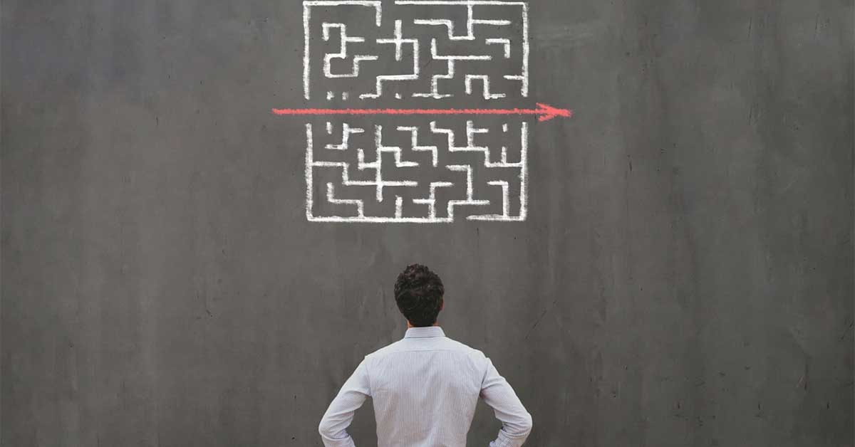 Male leader looking at a chalk drawn maze on a blackboard. A red line and arrow is through the centre of the maze.